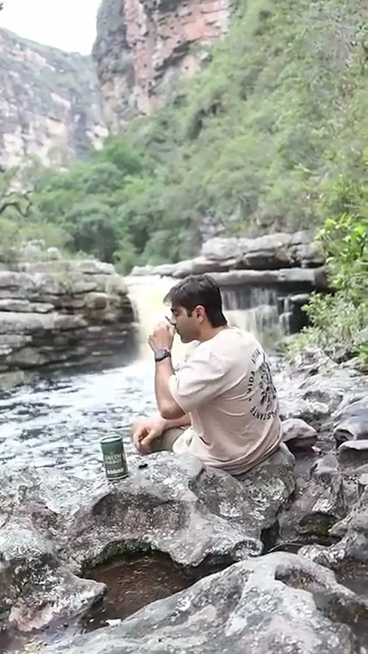 Homem sentado nas pedras de uma cachoeira prepara uma dose de Green Energy e toma.